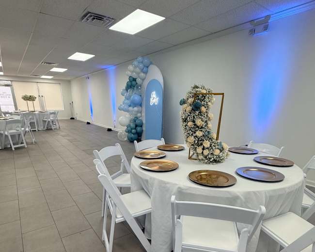 The image shows a decorated event space with a round table set for a gathering, featuring white chairs, blue and white balloons, and floral arrangements.