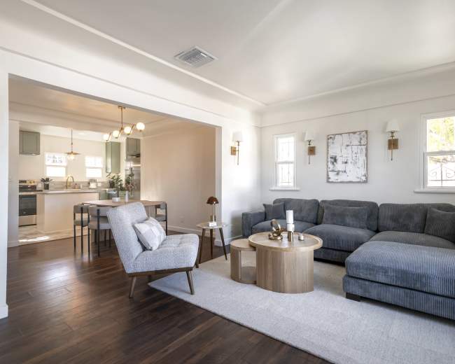 The image shows a modern living room with a gray sectional sofa, a round wooden coffee table, and a cozy armchair, adjacent to a dining area with a table and chairs.