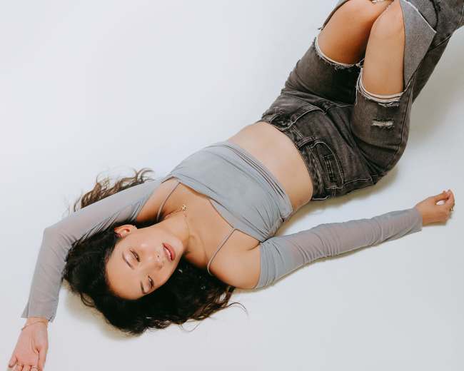 A person lies on a white surface wearing a gray crop top and distressed gray jeans.