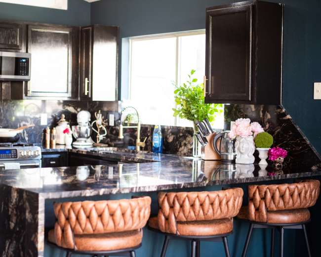 The kitchen features black cabinetry, a large dark marble countertop, and three brown leather bar stools.