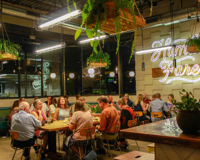 A group of people sits around wooden tables in a modern dining space decorated with hanging plants and a neon sign that says "Humble Fare."