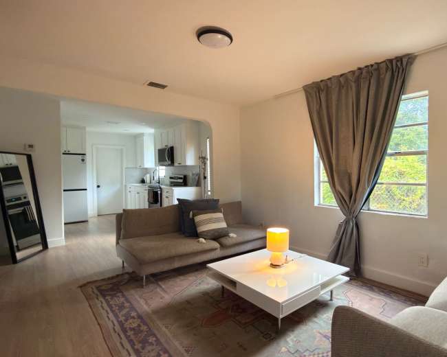 The image shows a modern living room with a couch, a coffee table, and large windows allowing natural light, connecting to a kitchen area in the background.