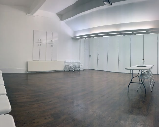 A spacious, empty room with wooden flooring, white walls, a folding table, and several white folding chairs arranged along one side.