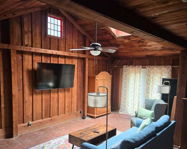 The interior of a rustic living space features a blue sofa, a wooden coffee table, a television mounted on the wall, and a ceiling fan, surrounded by wood-paneled walls and a patterned area rug.