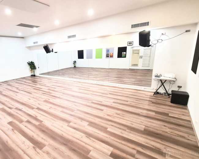 The image shows a spacious, empty dance studio with wooden flooring, mirrored walls, and minimal decor.