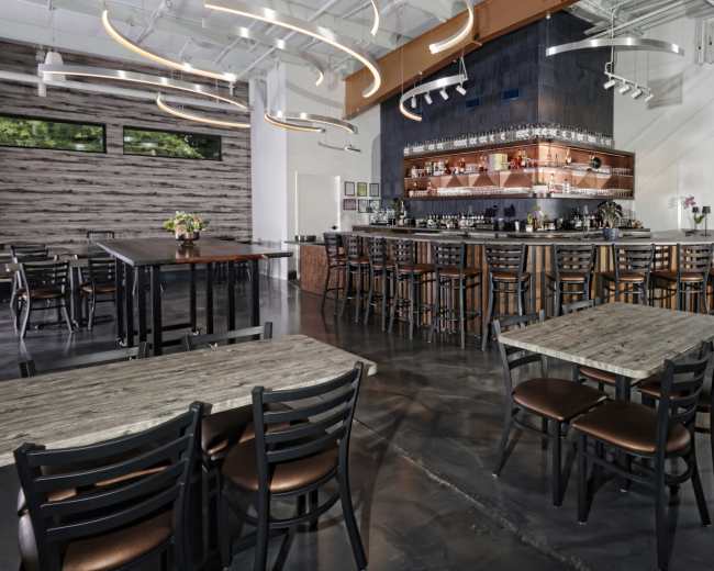 The image shows a spacious restaurant interior featuring a bar along one wall, wooden tables and chairs, and modern lighting fixtures.
