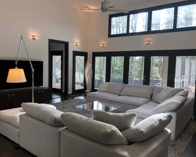 A modern living room with a large sectional sofa, a coffee table, and floor-to-ceiling windows overlooking a forested area.