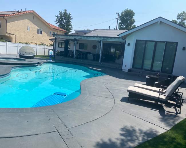 The image shows a landscaped backyard featuring a swimming pool, lounge chairs, and a modern house with large windows.
