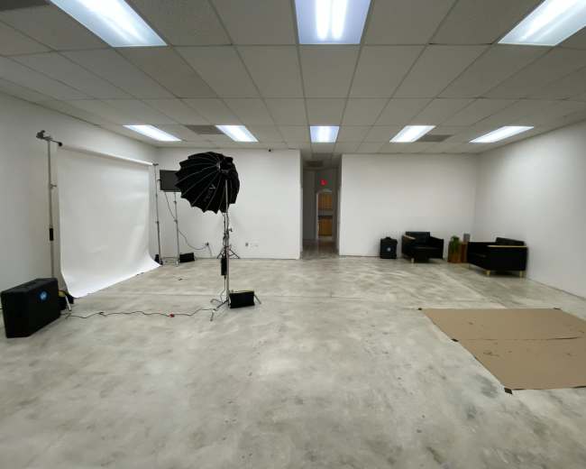 An empty studio space with gray concrete flooring, a backdrop setup with lighting equipment, and a few pieces of furniture along the walls.