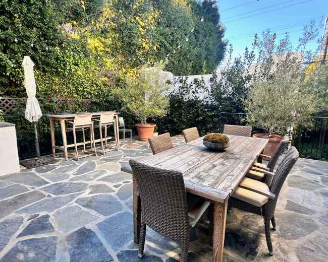 A patio features a large wooden table surrounded by wicker chairs, with a bar-height table and chairs set against a backdrop of greenery.