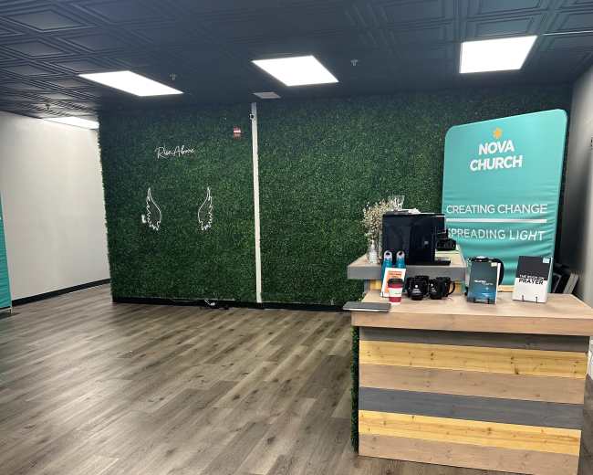 The interior of a modern church space features a wooden reception desk, a green accent wall with artificial grass, and a minimalistic layout with wooden flooring.