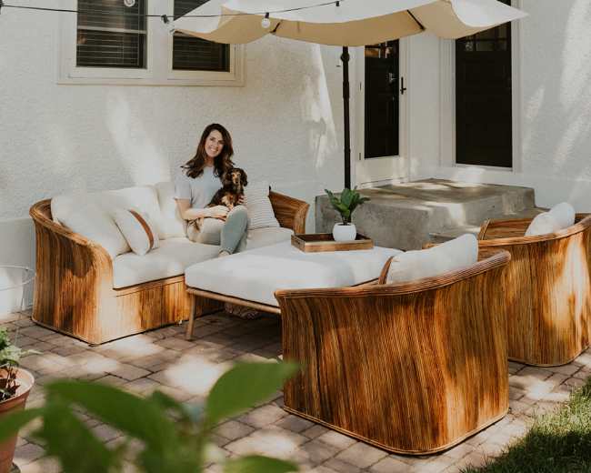 A woman sits on a wicker couch in a backyard area with a large umbrella and string lights above.