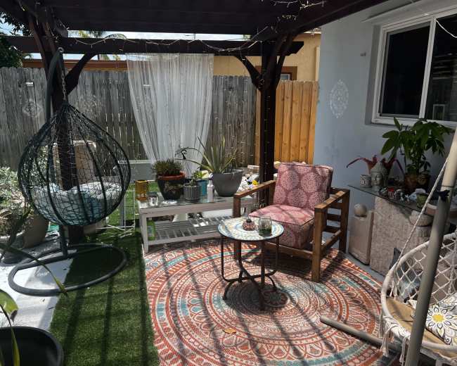 A shaded backyard nook features a hanging chair, a patterned rug, a small table, and various potted plants.