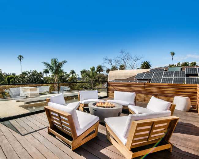 The image shows a rooftop seating area with four wooden chairs surrounding a fire pit and solar panels on a nearby structure under a clear blue sky.