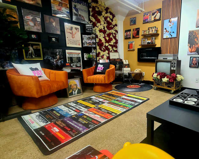 The room features two orange chairs, a black record player on the floor, a wall adorned with framed photographs and art, and a colorful area rug with a grid of album covers.