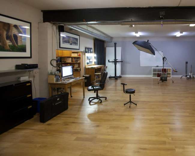 The image shows a spacious studio with wooden flooring, featuring a desk with a computer, a rolling chair, and a large red tool chest against the wall, along with various photography equipment set up in the background.