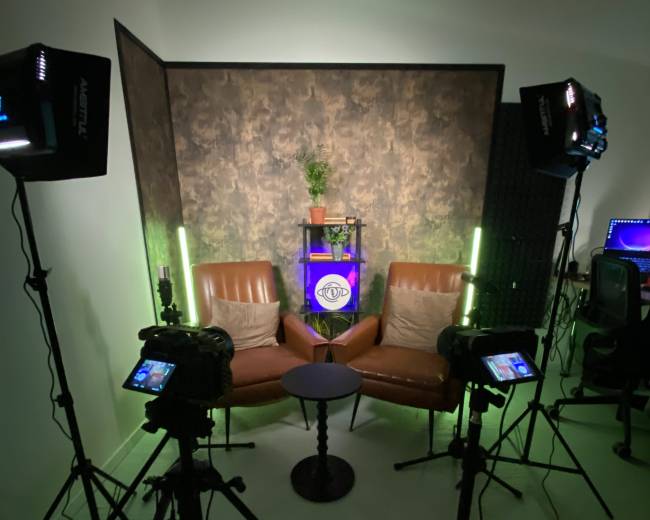 The image shows a small, well-lit studio setup featuring two brown chairs facing each other, a small round table in between, and various lighting equipment positioned around the scene.