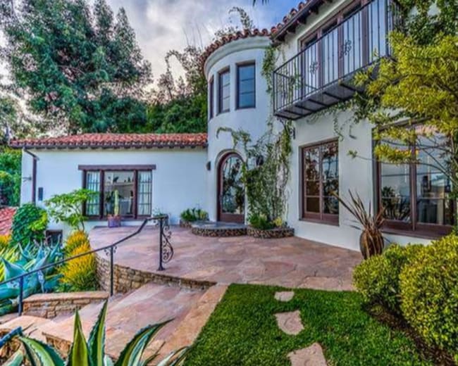 The image shows a two-story house with a stucco exterior, a tiled roof, and landscaped pathways leading to a front entrance surrounded by greenery.