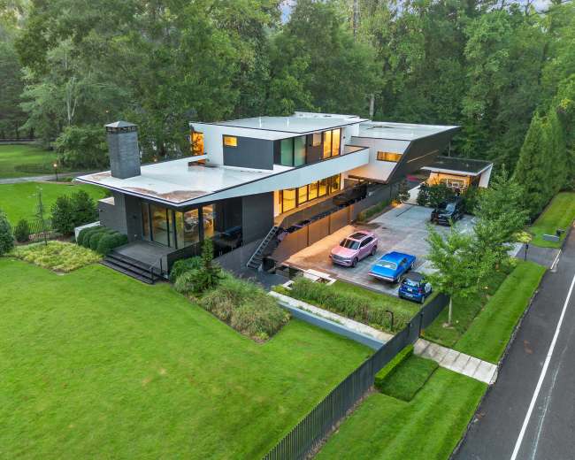 The image shows a modern, multi-level house with large windows and a unique angular roof, surrounded by a well-manicured lawn and several parked cars in the driveway.