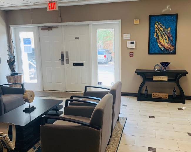 The image shows a waiting area featuring four chairs around a coffee table, with an art piece of an alligator on the wall, and a door leading outside.