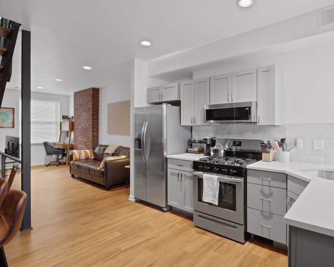A modern kitchen features stainless steel appliances, gray cabinetry, and an open layout that connects to a living area with a brown couch.
