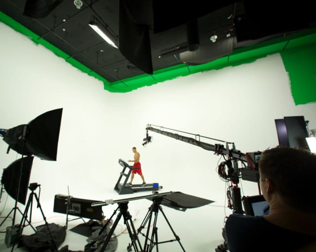 A person runs on a treadmill in a studio surrounded by professional lighting and filming equipment.