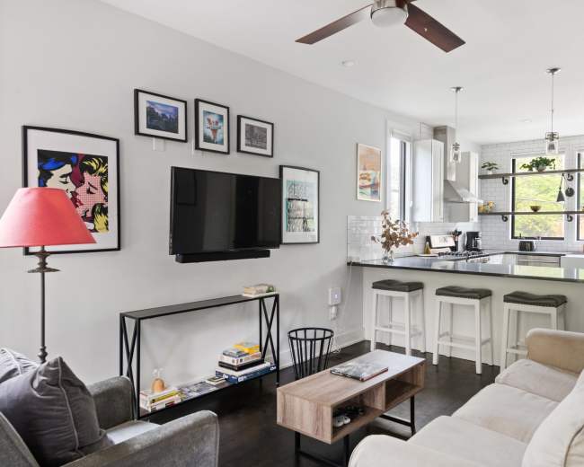 The image shows a modern living room and kitchen area with a mix of artwork on the walls, a television, and bar seating at the kitchen counter.