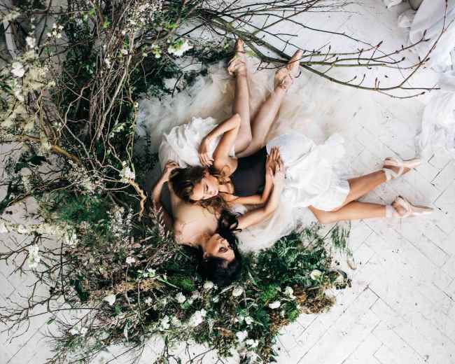 Two dancers in tutus rest on a bed of greenery and branches, surrounded by white flowers and soft fabric on a light floor.
