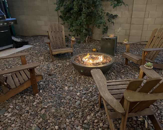 A circular fire pit surrounded by wooden chairs sits on a gravel surface in a cozy outdoor setting.