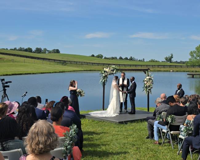 A bride and groom stand on a wooden platform beside a pond, while an officiant performs a wedding ceremony in front of guests seated on chairs.