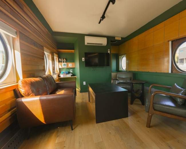 The image shows a small, modern living space with wooden walls, circular windows, a television mounted on the wall, and a seating arrangement consisting of two chairs and a leather sofa.