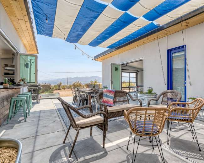A patio area with a striped awning, featuring several chairs and a small table, surrounded by potted plants and a view of mountains in the distance.