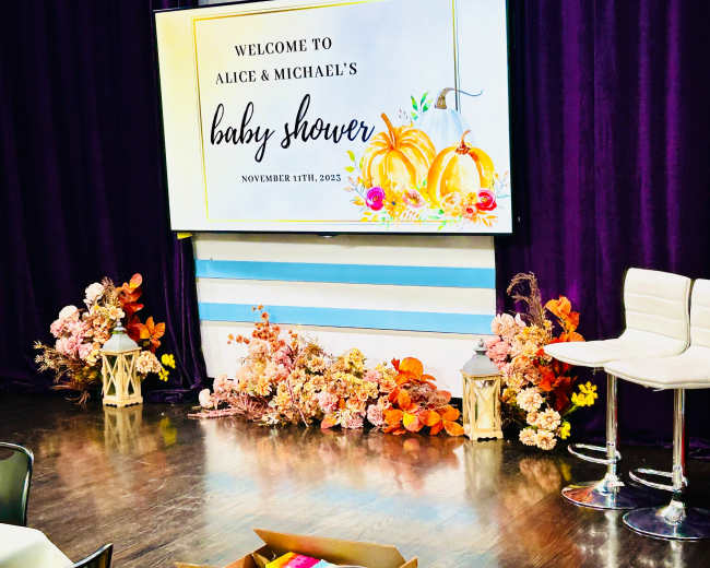 A decorated area for a baby shower, featuring a large display board with "Welcome to Alice & Michael's Baby Shower" and colorful floral arrangements at the base.