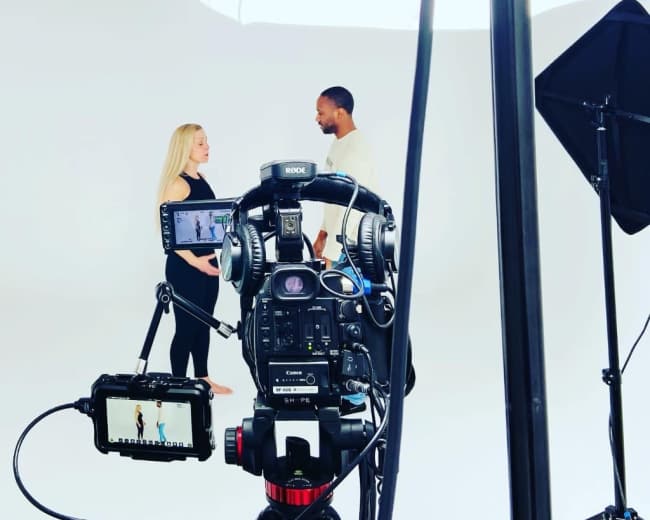 A camera set up on a tripod captures two individuals engaged in conversation in a well-lit studio against a plain white backdrop.