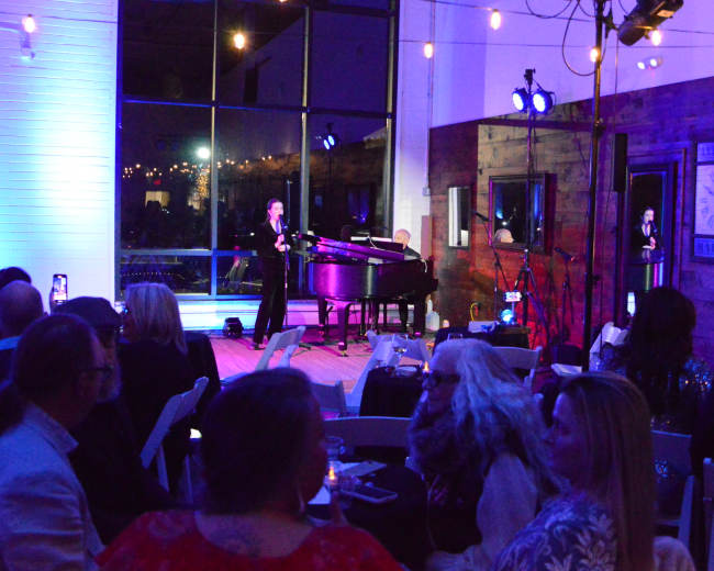 A pianist performs on stage in a dimly lit venue, while an audience watches and takes photos.