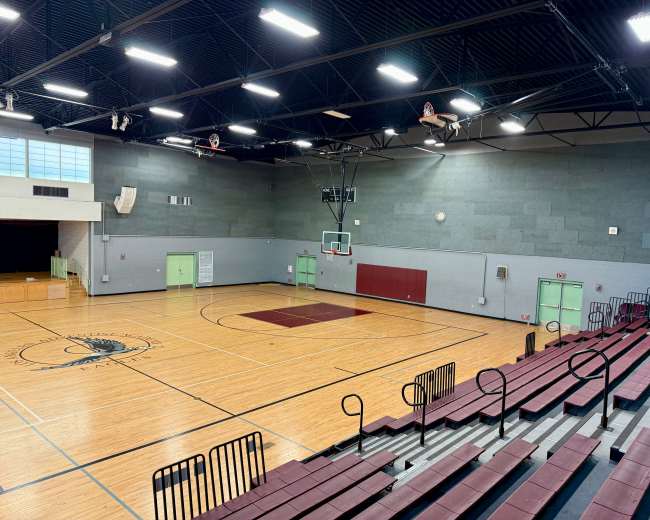 The image shows a spacious wooden gymnasium with bleachers, basketball hoops, and a polished court.