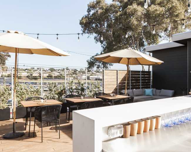 The image shows a rooftop terrace with tables, chairs, and umbrellas, overlooking a water view.