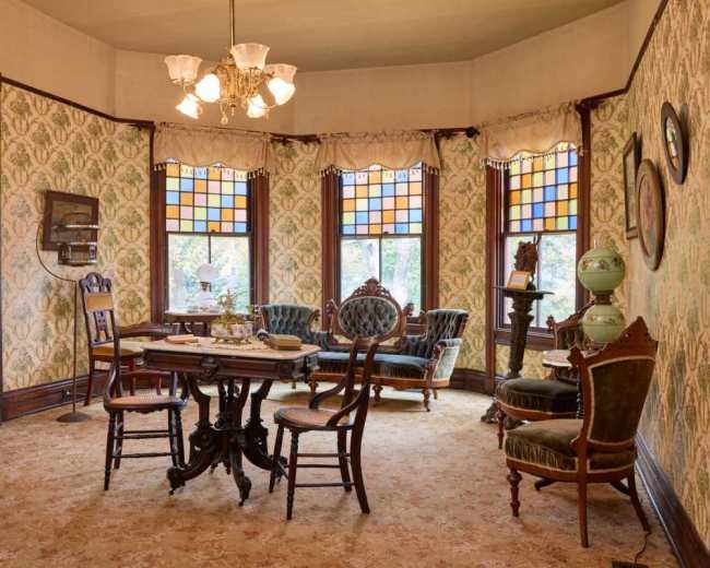 The image shows a vintage parlor room with patterned wallpaper, ornate furniture, and colorful stained glass windows.