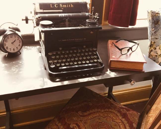 The scene features an antique LC Smith typewriter on a desk alongside a vintage clock, a pair of glasses, and a book, with sunlight filtering through a window draped in red curtains.