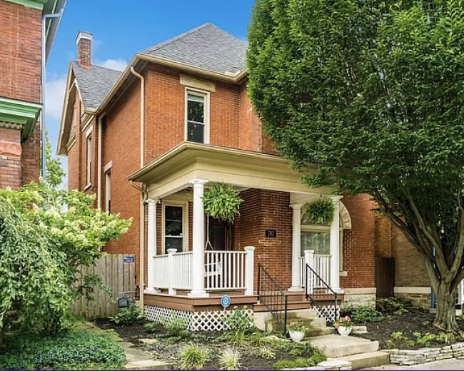 The image shows a red brick Victorian-style house with a porch, surrounded by greenery and well-maintained landscaping.