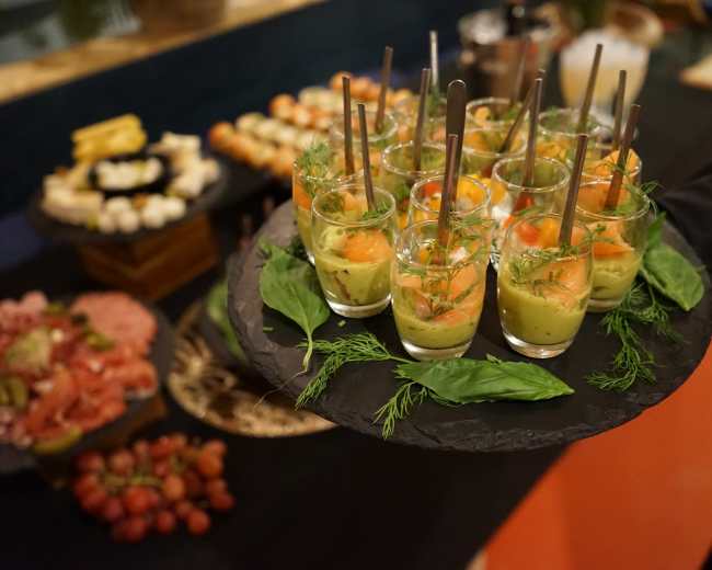 A server holds a black tray with glasses of layered green and orange appetizers, garnished with herbs, alongside a spread of various finger foods in the background.