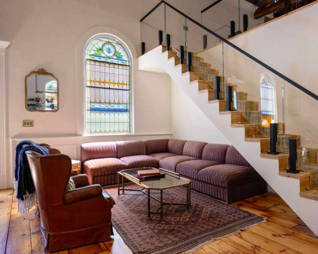 The image shows a stylish living room with a curved sectional sofa, a wooden coffee table, and a staircase featuring glass railings and a stained glass window.