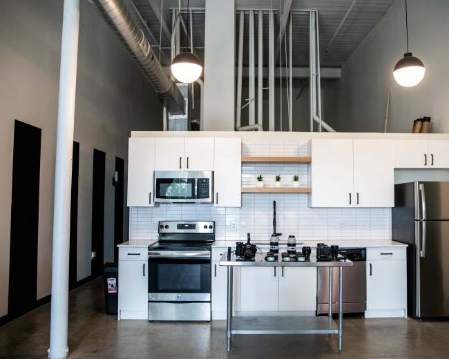 The image shows a modern kitchen featuring white cabinets, stainless steel appliances, a central island, and a high ceiling with visible piping.