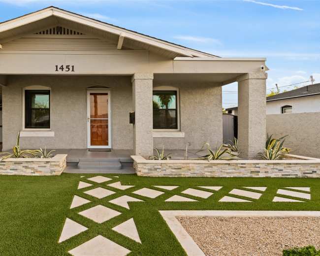 The image shows a modern single-story house with a stone-accented facade, manicured lawn, and geometric pathway leading to the front door.