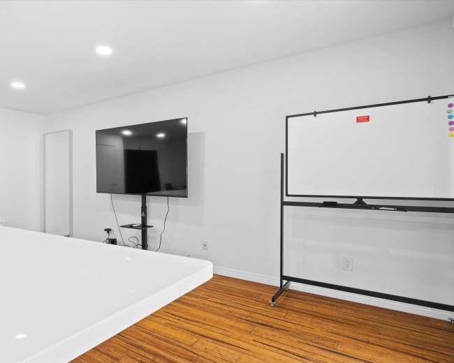 The image shows a modern meeting room featuring a large flat-screen TV mounted on the wall and a whiteboard on a movable stand, with white walls and a wooden floor.