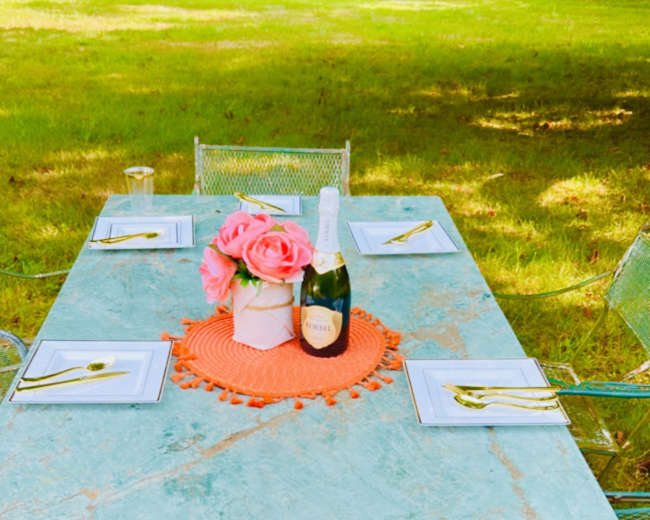 A marble table is set with gold utensils and white plates, adorned with a floral centerpiece and a bottle of champagne, surrounded by chairs on a grassy area.