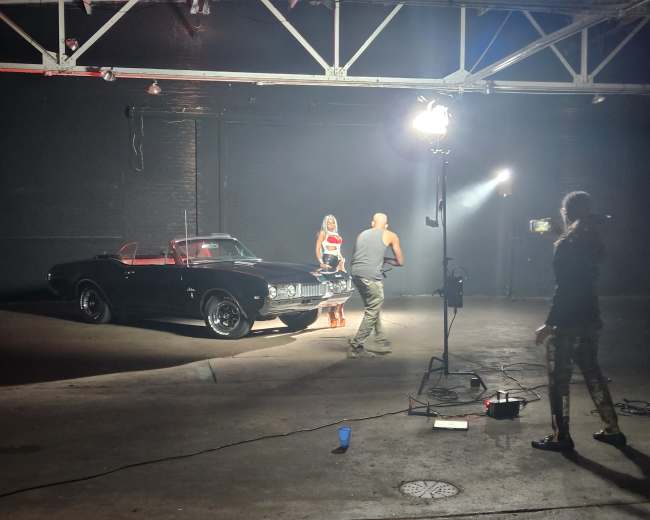 A classic black convertible car is positioned in a dimly lit industrial space while a person interacts with a model in front of it.