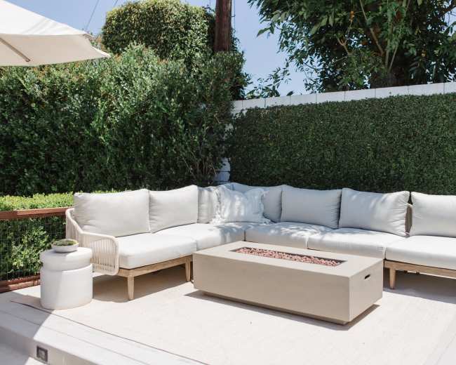 A modern outdoor seating area features a light-colored sectional sofa and a rectangular fire pit surrounded by greenery.