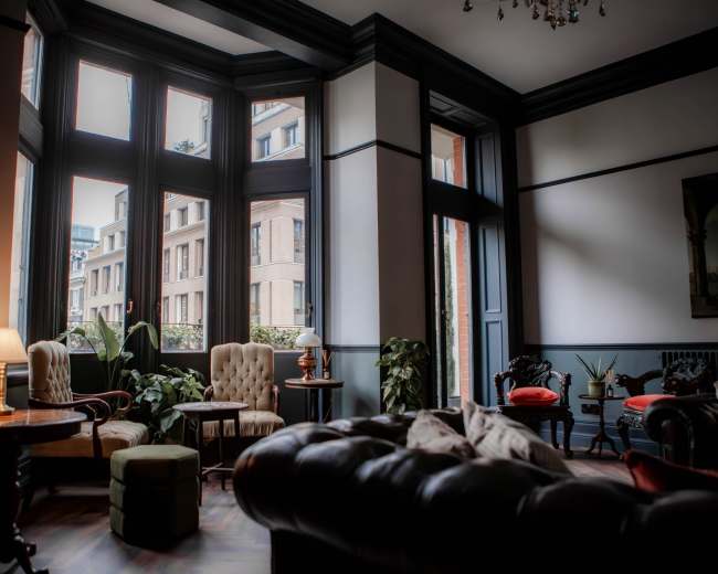 The image shows a warmly lit lounge with several seating options, large windows, and indoor plants overlooking a cityscape.