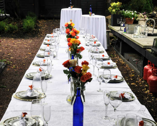 A long, elegantly set outdoor dining table is adorned with floral centerpieces and glassware, ready for a meal.